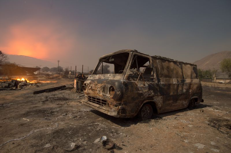 a  burned van rests in a lot while a wildfire glows on the horizon
