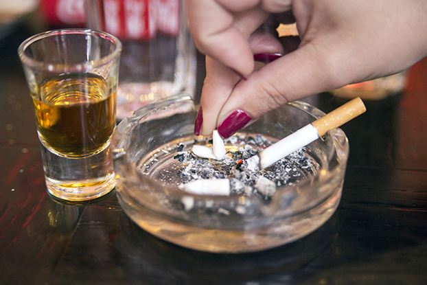 Full shot glass with woman's hand putting out cigarette in full ashtray